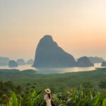 As the sun sets, a figure stands in contemplation before the majestic mountains rising from the calm waters, surrounded by lush greenery and serenity. Phangnga Bay Thailand