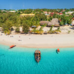 Aerial view of boats on tropical sea coast with sandy beach at sunny day. Summer holiday on Indian Ocean, Zanzibar, Africa. Landscape with boat, palm trees, transparent blue water, hotels. Top view