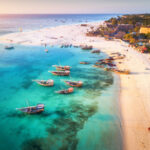 Aerial view of the fishing boats on tropical sea coast with sandy beach at sunset. Summer holiday on Indian Ocean, Zanzibar, Africa. Landscape with boat, buildings, transparent blue water. Top view