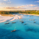 Aerial view of the fishing boats on tropical sea coast with sandy beach at sunset. Summer travel in Zanzibar, Africa. Top view of boats, yachts, green palm trees, clear blue water, colorful sky