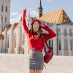 Excited caucasian girl in glamorous outfit posing emotionally near old beautiful palace. Amazing brunette lady in glasses holding backpack and dancing in front of white castle.