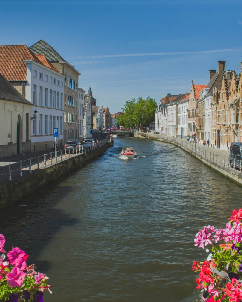 Charming view of Bruges' canal with vibrant flowers, historical buildings, and a tourist boat on a sunny day.