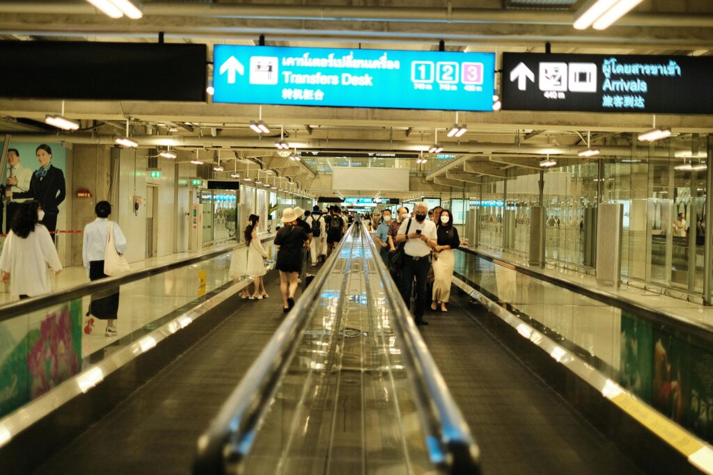 Passengers walk on a travelator in an airport arrival area, surrounded by signs and bright lighting.
