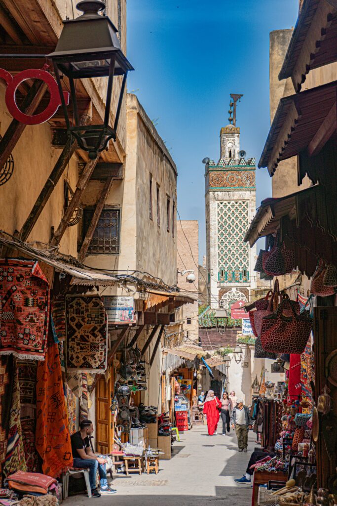 Bustling street in Fes, Morocco with shops, textiles, and a minaret in view.