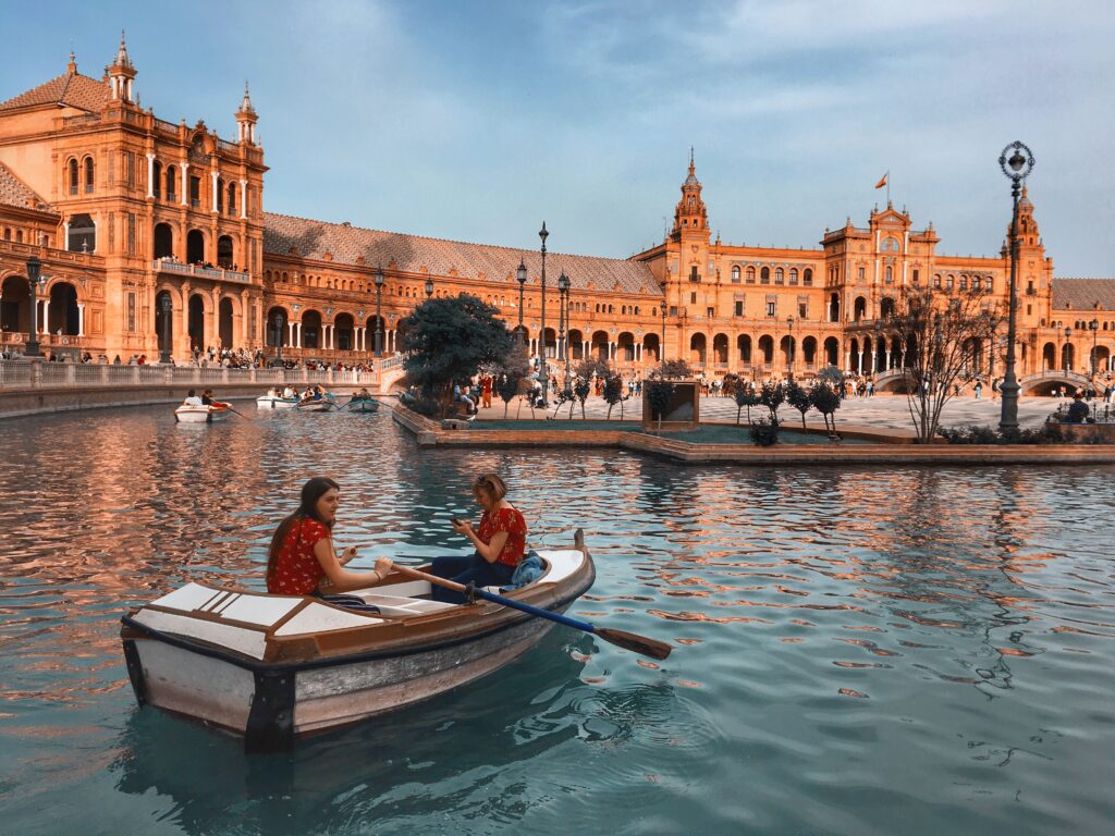 Two women enjoy a boat ride on the canal at Plaza de España in Seville, Spain, surrounded by beautiful architecture.